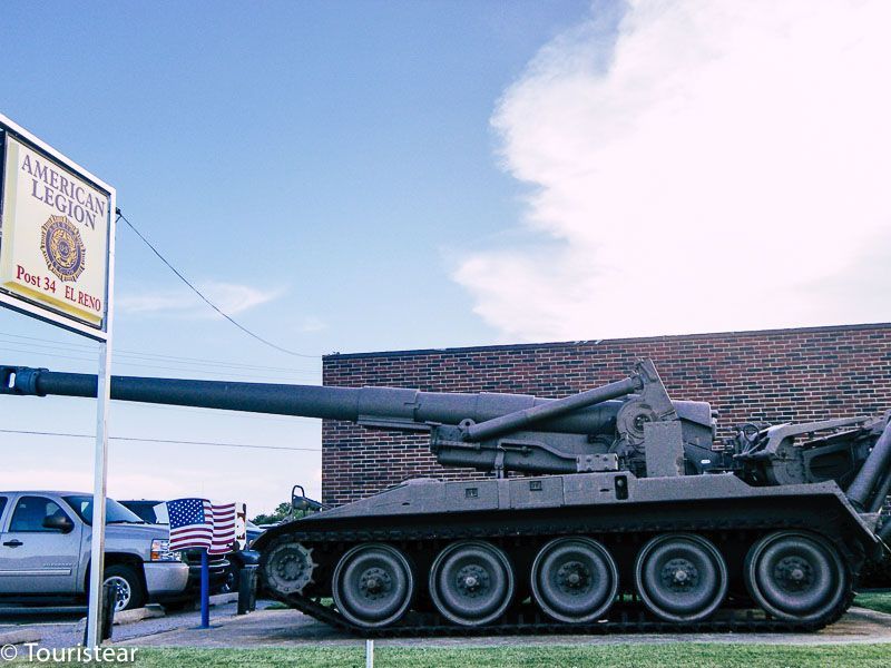 A tank on display beside a bricked wall building under bright blue skies with white clouds.