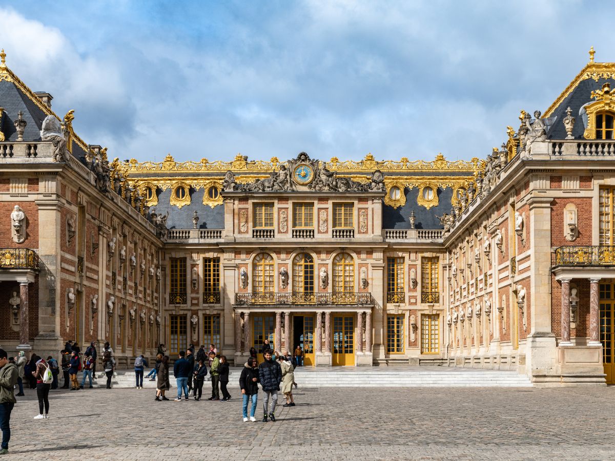 The front of the Palace Versailles filled with people taking photos and walking during a day trip from Paris